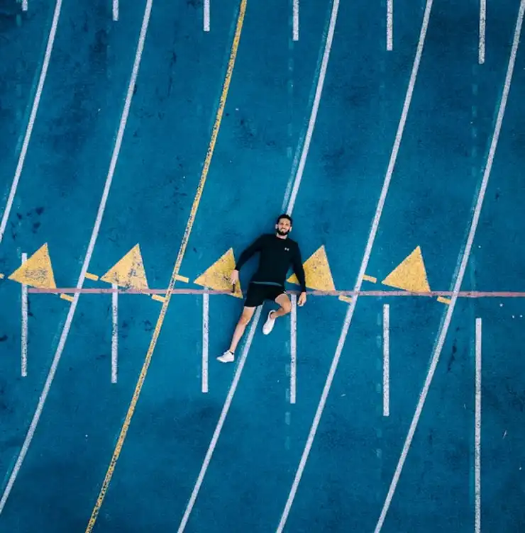 Um homem em roupa de desporto está deitado numa pista de corrida de um azul vibrante, marcada por linhas brancas e triângulos amarelos. A cena transmite uma impressão de relaxamento e contraste.