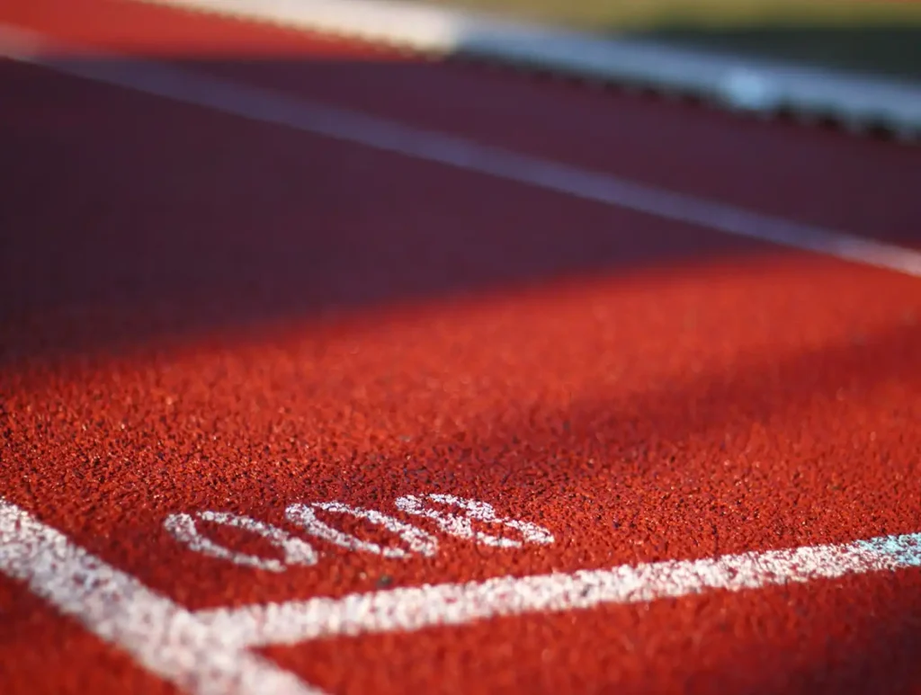 Close-up de uma pista de atletismo vermelha marcada com o número "800" em branco, destacando a superfície texturizada e as linhas de demarcação nítidas sob a suave luz do sol.