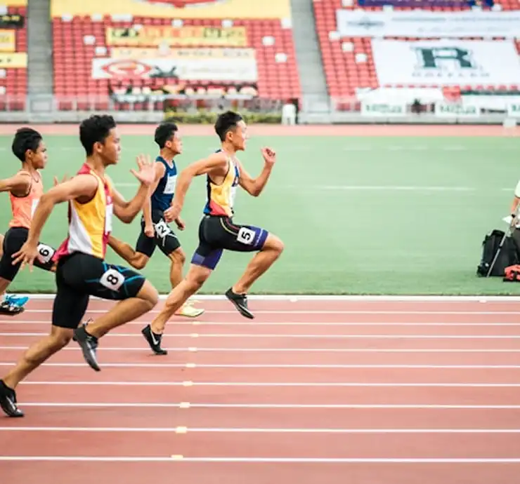 Cinco atletas masculinos correm a toda velocidade numa pista vermelha num estádio, com concentração e determinação. Ao fundo, as arquibancadas estão adornadas com faixas coloridas.