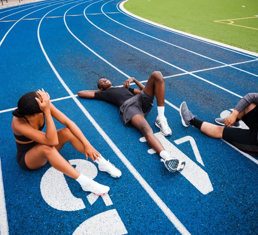Três atletas em roupa de desporto descansam numa pista de corrida de um azul brilhante. Dois deles estão sentados, com a cabeça baixa, enquanto o terceiro está deitado de costas, parecendo exausto mas satisfeito.