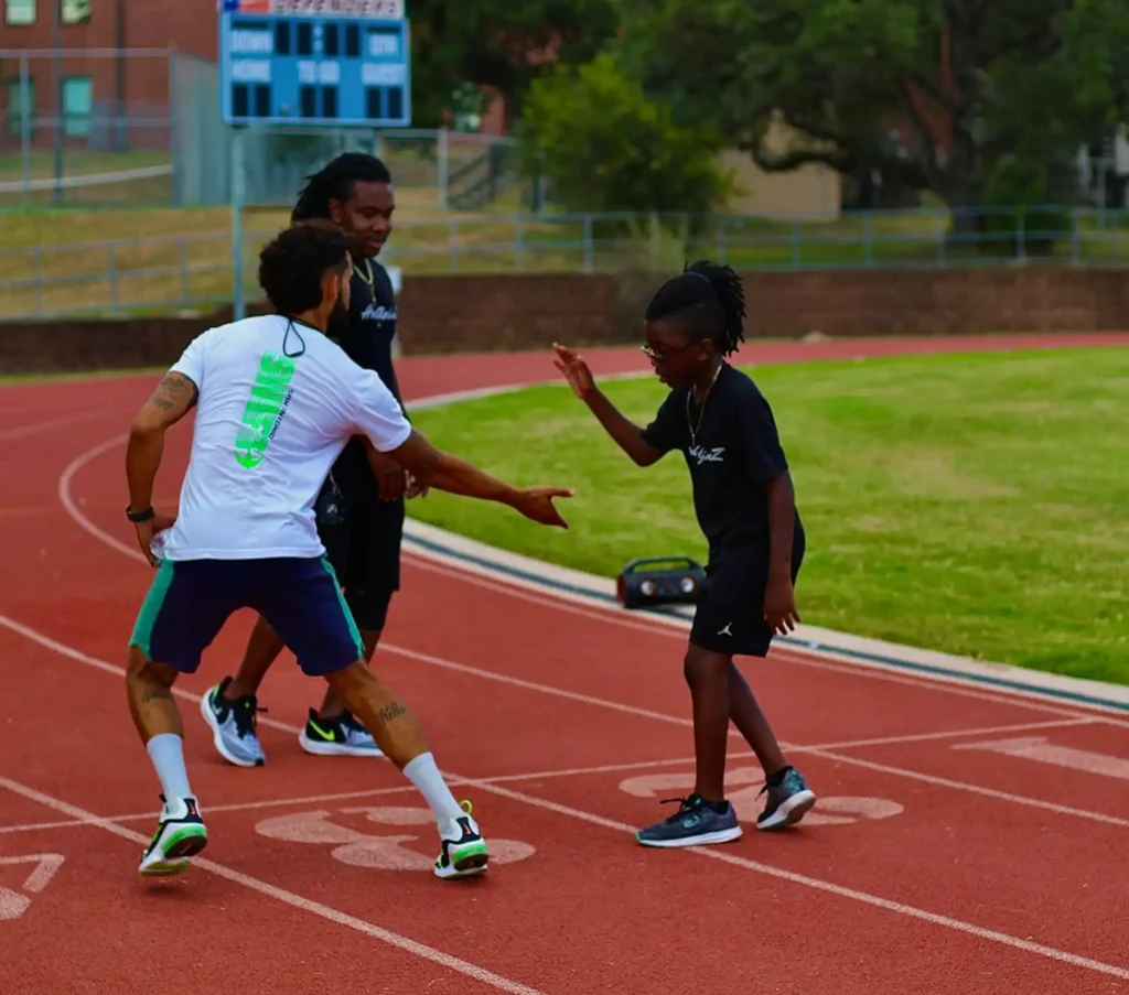 Três pessoas numa pista celebram uma corrida bem-sucedida. Um homem bate palmas com um garoto sorridente, num gesto de solidariedade e encorajamento. Um placar é visível ao fundo.