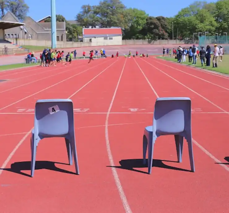 Duas cadeiras cinzas colocadas numa pista de atletismo vermelha estão viradas para uma multidão distante. Sob um céu azul límpido, a cena transmite uma sensação de expectativa e serenidade.