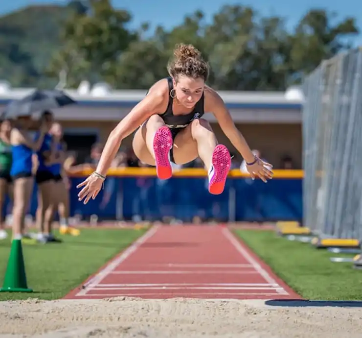 Uma atleta em pleno salto em distância, usando tênis rosa vibrante, com uma expressão concentrada. Dia ensolarado, pista e espectadores ao fundo.